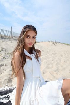 a beautiful young woman sitting on top of a beach next to the ocean wearing a white dress