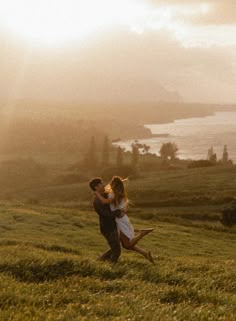 a man holding a woman on top of a lush green field next to the ocean