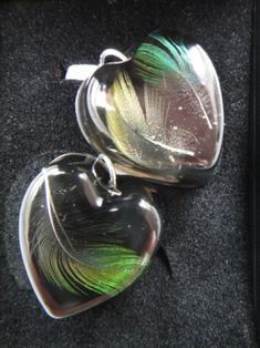 two silver heart shaped pendants with green feathers on them sitting in a black box