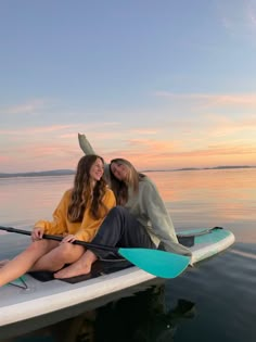 two women sitting on a surfboard in the water at sunset or dawn with their arms around each other