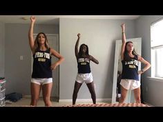 three girls in cheerleader shirts are standing on a bed and one girl is holding her arms up