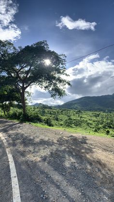 the sun shines brightly through the clouds above a tree on a dirt road with mountains in the background