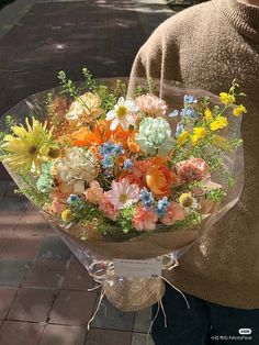 a woman holding a bouquet of flowers on the street in front of her face and wearing a brown sweater