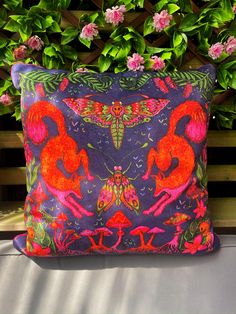 a purple pillow with pink flamingos on it sitting in front of some bushes and flowers