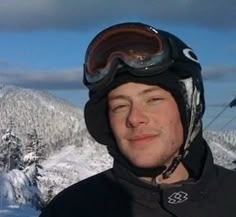 a man with ski goggles on standing in front of snow covered mountains and trees