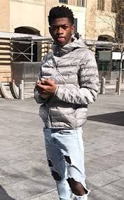 a young man standing on the sidewalk looking at his cell phone while wearing ripped jeans and a puffy jacket