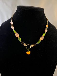 a gold tone necklace decorated with orange and brown beads with green leaf beads and gold tone spacers. A gold tone hoop connects the two sides of the necklace with a small pumpkin hanging from the bottom of the hoop. Pumpkin Necklace, Orange Decor, Small Pumpkins, Gold Tone Necklace, Green Leaves, Fall Pumpkins, Gold Tones, Two By Two, Beaded Necklace