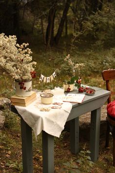 a table with flowers and books on it in the middle of a wooded area next to a wooden chair