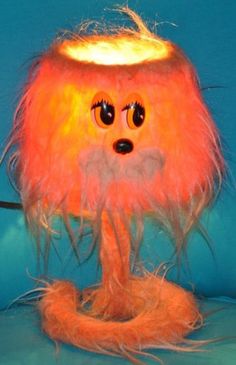 an orange furry lamp with eyes and long hair on it's head, sitting on a blue surface