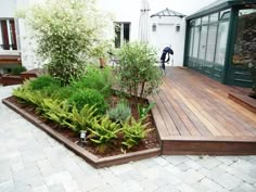 a wooden deck surrounded by greenery and plants in front of a white building with green doors