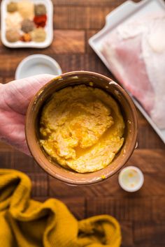 a person holding a mug filled with yellow food on top of a wooden table next to bowls