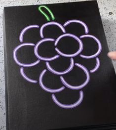 a hand is holding up a black canvas with purple and green grapes drawn on it