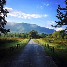 an empty country road with mountains in the distance and trees on either side that are fenced off