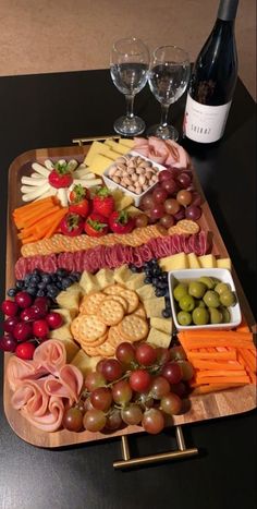 a wine bottle and two glasses on a table with a platter of cheese, crackers, grapes, strawberries, meats and vegetables