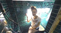 a woman sitting on top of a toilet seat in a green tiled room with sunlight coming through the window