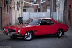 a red car parked on the side of a road next to tall brick buildings in an alleyway