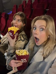 two women eating popcorn while sitting in the theater seats with their mouths open and looking surprised