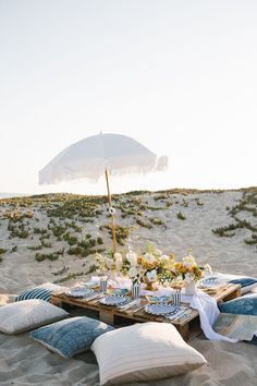 a table set up on the beach with pillows and an umbrella in the sand for dinner