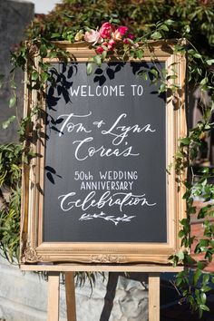 a sign that is sitting in front of a building with flowers and greenery on it