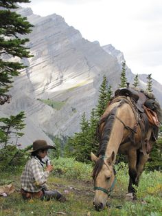 a man kneeling down next to a horse on top of a grass covered hillside with mountains in the background