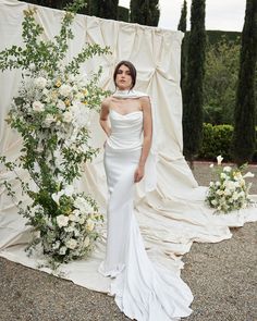 a woman in a white dress standing next to a flower covered backdrop with roses and greenery