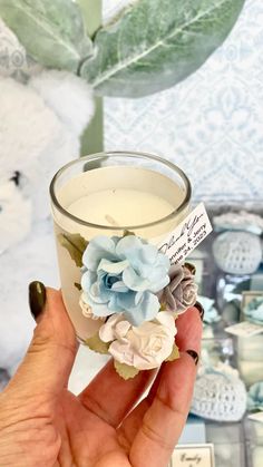 a hand holding a small glass candle with flowers on the inside and label in front of it