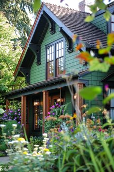 a green house with lots of flowers in the front yard and trees around it on a sunny day