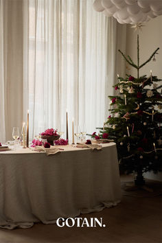 A Christmas-themed dining room with a festive table setting, a glowing Christmas tree, and sheer linen curtains in sand filtering soft natural light.