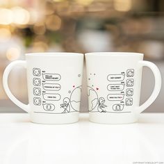 two white coffee mugs sitting on top of a table with stickers drawn on them