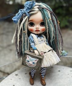 a doll with dreadlocks is standing on a rock and holding a small box