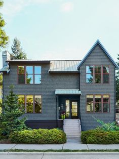 a gray house with red trim on the front and side windows, bushes and trees