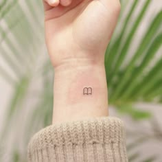 a woman's wrist tattoo with the letter m on her left hand, and a green plant in the background