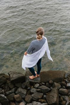 a woman standing on rocks near the water with her arms wrapped around her body and looking at the water