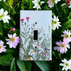 a light switch cover with flowers painted on it in front of green leaves and pink daisies