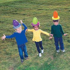 three children wearing colorful hats standing in the grass with their arms out to each other