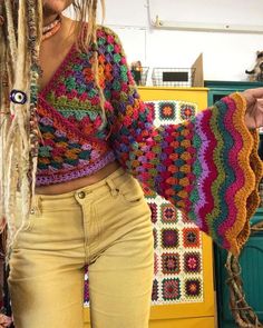 a woman with dreadlocks standing in front of a yellow cabinet wearing a multicolored crochet sweater