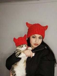 a woman holding a cat in her arms and wearing a red knitted devil hat