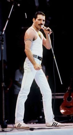 a man standing on stage with a microphone in his hand and wearing white pants,