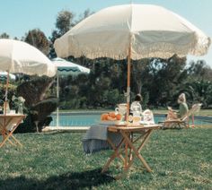 two people sitting at a table with an umbrella over them in front of a pool