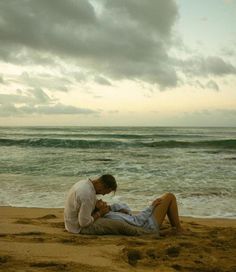 a man and woman sitting on top of a sandy beach next to the ocean under a cloudy sky