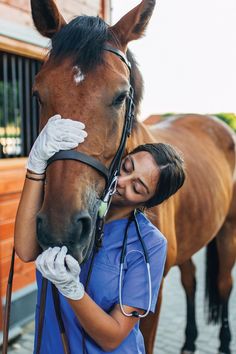 a woman in scrubs is petting a brown horse's head while standing next to a stable