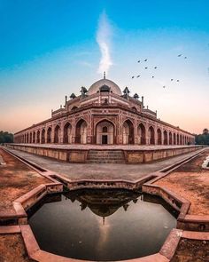 an old building with a pond in front of it and birds flying around the area