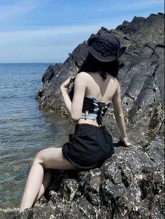 a woman sitting on top of a rock next to the ocean wearing a black hat