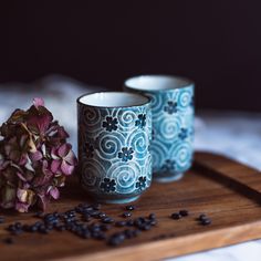 two coffee mugs sitting on top of a wooden cutting board next to dried flowers