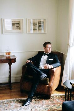 a man in a tuxedo sitting on a chair with his legs crossed, looking off to the side