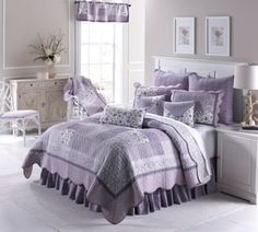 a bed with purple bedspread and pillows in a white room next to a window