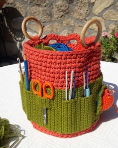 a knitted basket with scissors and knitting needles in it on top of a table