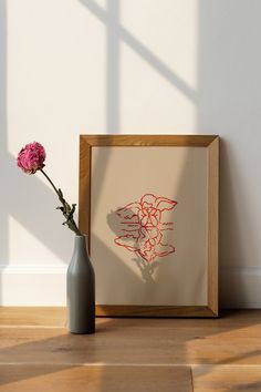 a vase with a pink flower in it next to a framed drawing on the wall