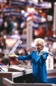 an older woman standing at a podium with her arms outstretched in front of a crowd