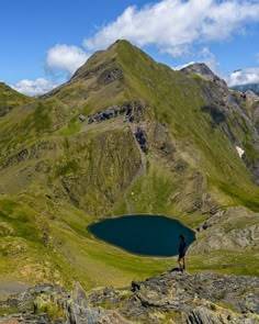 a man standing on top of a lush green hillside next to a blue lake in the mountains